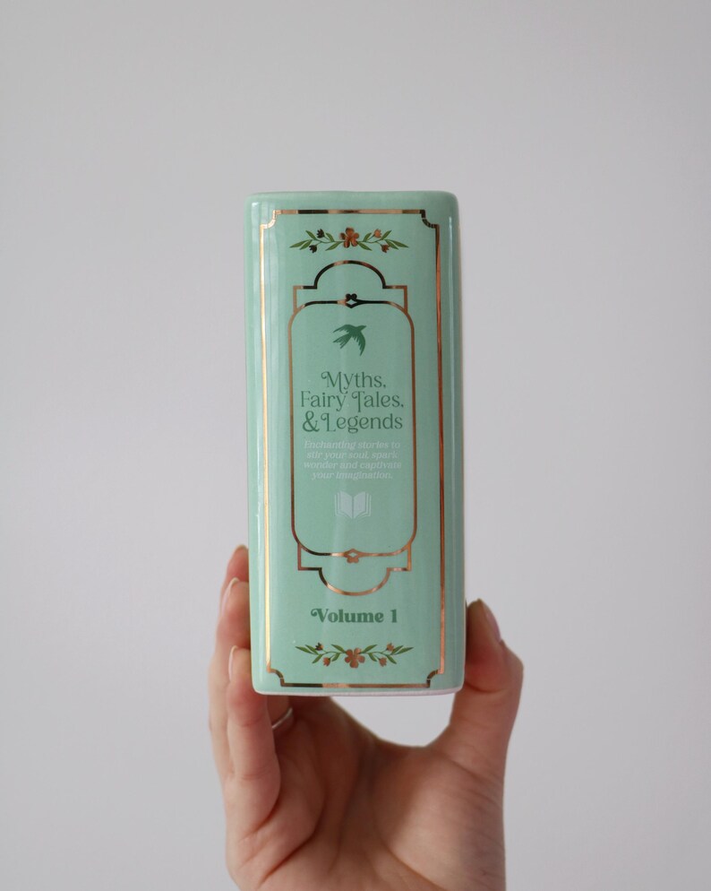 May include: A rectangular, mint-green ceramic container with gold trim and the text "Myths, Fairy Tales, & Legends, Volume 1." The design includes floral and bird motifs, suggesting a literary theme.