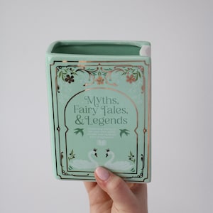 May include: A mint green ceramic book-shaped vase with gold trim and the words "Myths, Fairy Tales, & Legends." The design includes floral accents and two swans. The vase is held by a person.