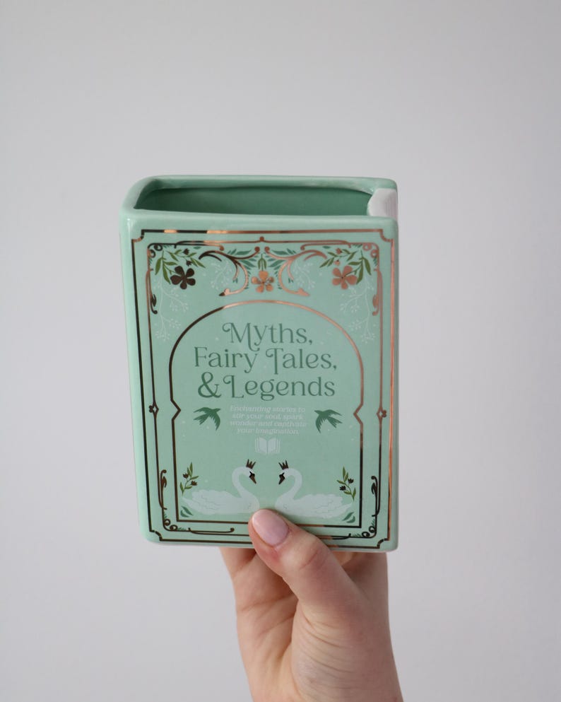 May include: A mint green ceramic book-shaped vase with gold trim and the words "Myths, Fairy Tales, & Legends." The design includes floral accents and two swans. The vase is held by a person.