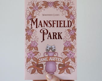 May include: A pink paperback book titled "Mansfield Park" by Jane Austen. The cover features floral and ribbon designs in shades of pink and brown, with a decorative vase and theater masks.