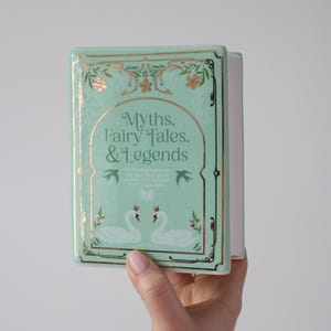 May include: A mint green book titled "Myths, Fairy Tales, & Legends" with gold accents and a decorative border. The cover features two swans and floral designs. The book is held in a hand.