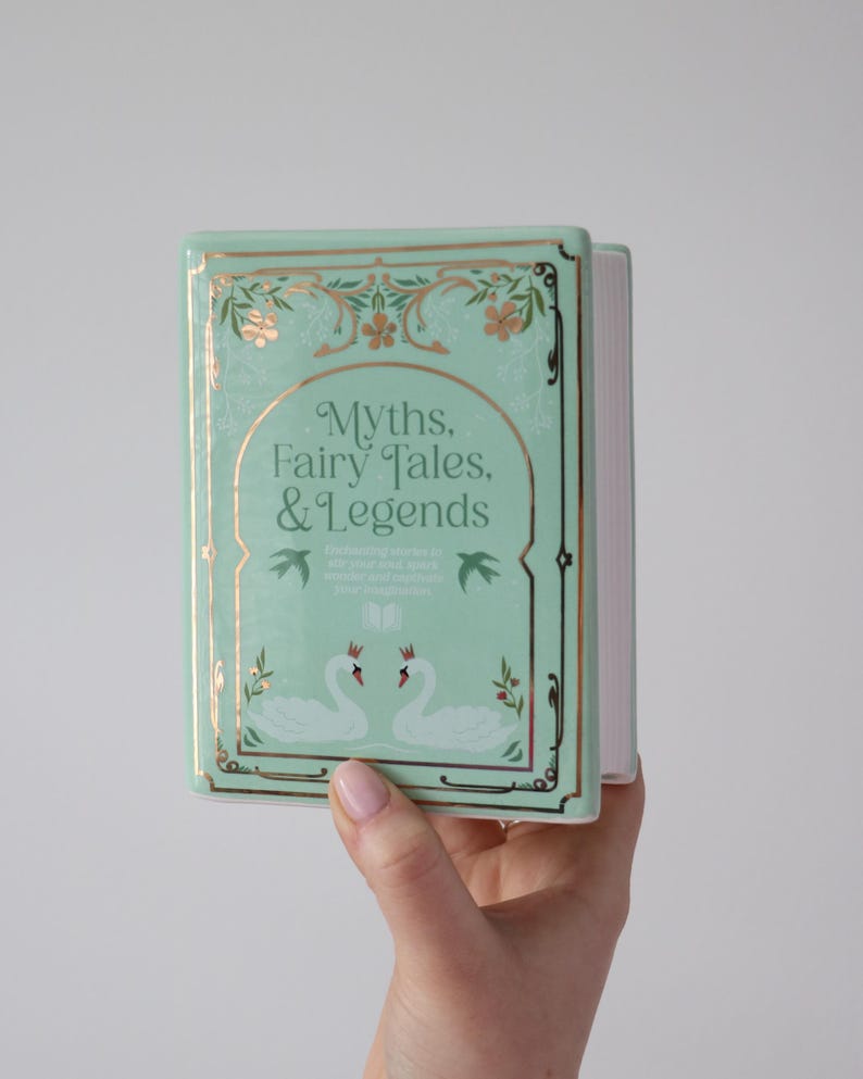 May include: A mint green book titled "Myths, Fairy Tales, & Legends" with gold accents and a decorative border. The cover features two swans and floral designs. The book is held in a hand.