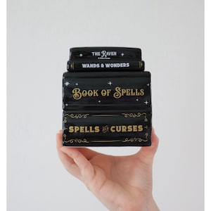 May include: A black ceramic book-shaped box set with gold lettering. Titles include "The Raven," "Wands & Wonders," "Book of Spells," and "Spells and Curses." The set is held in a hand against a white backdrop.