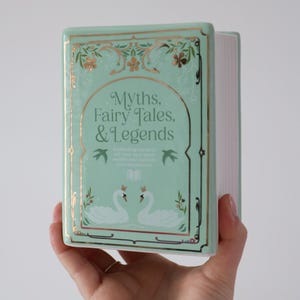 May include: A mint green ceramic book with gold accents and the title "Myths, Fairy Tales, & Legends." The book features illustrations of swans and floral designs. The book is held in a hand.