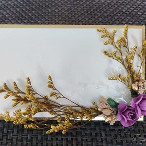 May include: A white card with a gold border, decorated with dried flowers and purple silk flowers. The card is on a woven brown background.