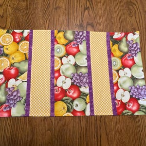 May include: A yellow and white table runner with a fruit pattern. The runner has purple stripes and a yellow background with a white polka dot pattern.