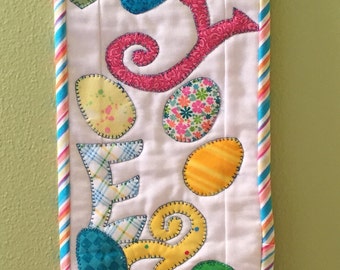 May include: A quilted Easter banner with the words "Happy Easter" in colorful, patterned fabric letters. The banner includes appliqued Easter eggs and a striped border, perfect for spring decor.