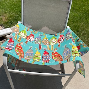 May include: A turquoise tablecloth with a colorful bird pattern. The birds are red, yellow, green, blue, and white. The tablecloth is draped over a white metal chair.