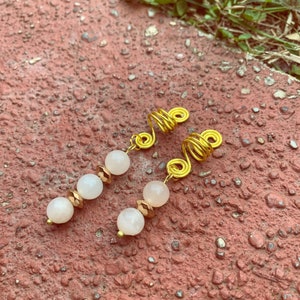 May include: Two gold-colored hair spirals with dangling beads. Each spiral has three light pink beads and two gold-colored faceted beads. The hair accessories are displayed on a red brick surface.