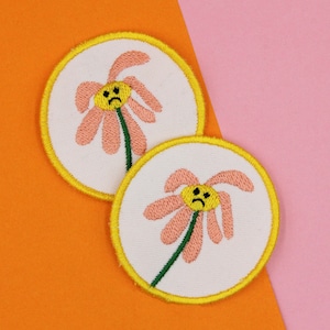 Peut inclure: Deux écussons brodés avec un motif de fleur au visage triste. La fleur a des pétales roses, un centre jaune et une tige verte. Les écussons sont circulaires avec un fond blanc et une bordure jaune. Le fond est orange et rose.