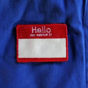 Peut inclure: Un badge nominatif rouge et blanc avec les mots "Hello my name is" en lettres blanches. Le badge est rectangulaire et cousu sur un tissu bleu. Un bouton bleu est visible.