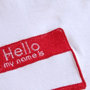 Peut inclure: Tissu blanc avec une étiquette nominative rouge brodée. L'étiquette indique "Hello my name is" en fil blanc. L'étiquette nominative est rectangulaire avec des coins arrondis.