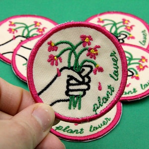 Puede incluir: Parche circular bordado con fondo crema, que presenta una mano sosteniendo un ramo de flores rosas con tallos verdes. El parche tiene un borde rosa y las palabras "plant lover" en verde.