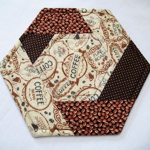 May include: Hexagon-shaped coffee-themed quilted coaster with a brown, beige, and white color scheme. The coaster features a repeating pattern of coffee beans, coffee words, and coffee-themed illustrations.