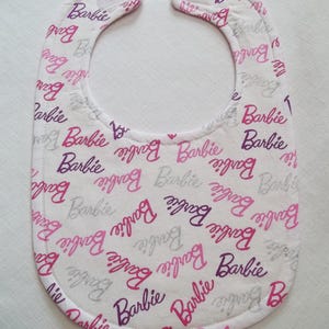 May include: White baby bib with a repeating pattern of the word "Barbie" in shades of pink, purple, and gray. The bib has a rounded shape with a curved neckline and is edged with a stitched border.