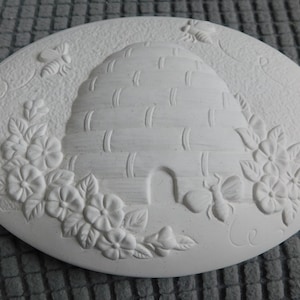 May include: Oval, off-white ceramic plaque featuring a raised relief design of a beehive, flowers, and bees. The beehive is in the center, surrounded by floral and bee motifs. The plaque is approximately 8 inches in diameter.