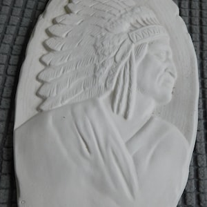 May include: A white plaster relief sculpture depicting a Native American chief in profile, wearing a feathered headdress. The sculpture is oval-shaped and has a textured surface, with details of the face and clothing.