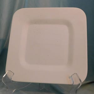 May include: A square white plate with rounded corners and a smooth, matte finish. The plate features a subtle, light gray speckled pattern across its surface and is displayed upright on a clear, transparent plastic stand for viewing.