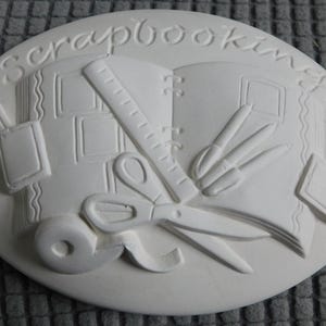 May include: A white oval-shaped plaque with the word "scrapbooking" embossed at the top. The plaque features embossed images of a scrapbook, scissors, a ruler, pens, and tape. The design is a craft-themed decorative item.