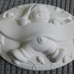 May include: A white, oval-shaped ceramic Christmas ornament featuring a smiling Santa Claus figure holding a banner. The ornament has a relief design with details like a hat, lights, and a decorative border.