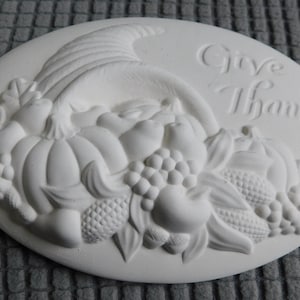 May include: An oval, unpainted plaster relief featuring a cornucopia overflowing with pumpkins, grapes, and other harvest fruits. The words "Give Thanks" are embossed on the top right. A decorative piece for fall.