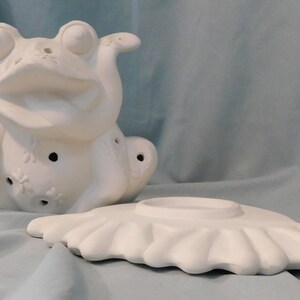May include: A white ceramic frog figurine with a hollow body and cut-out dragonfly patterns, accompanied by a separate white ceramic base shaped like a large flower petal. The frog has large eyes and a wide mouth, designed for decorative use.