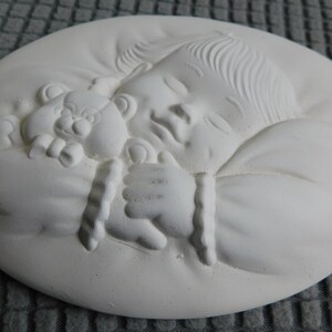 以下が含まれることがあります： 白い円形のレリーフ彫刻で、テディベアを抱きしめて眠っている赤ちゃんが描かれています。赤ちゃんは柔らかく丸みを帯びた形で、顔の特徴や髪の毛が詳細に表現されています。テディベアも詳細に描かれています。