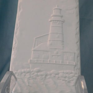 May include: A rectangular white ceramic plaque featuring a raised relief design of a lighthouse situated on a rocky shoreline. The plaque has two small holes at the top for hanging and displays a detailed lighthouse structure with a lantern room.