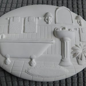 May include: A white, oval-shaped wall plaque featuring a bathroom scene. The relief carving depicts a bathtub, sink, towels, and a potted plant. The details include a rug, lamps, and a mirror.
