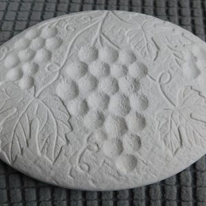 May include: A round, off-white decorative stone with a grape and leaf design. The design is embossed on the surface, featuring clusters of grapes and detailed leaves. The stone is approximately 8 inches in diameter.