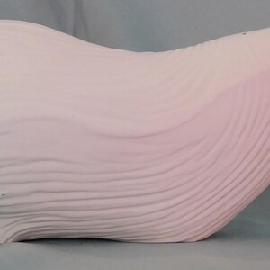May include: A matte white ceramic seashell sculpture featuring a series of horizontal, wavy, and raised ridges that follow the natural curvature of the shell. The object has a smooth, uniform finish and a distinct, elongated, and organic shape.