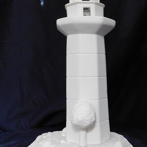 May include: A white ceramic lighthouse figurine. The lighthouse has a detailed design, including a tower with rectangular windows and a small, round tree-like embellishment. It sits on a textured base, perfect for home decor.