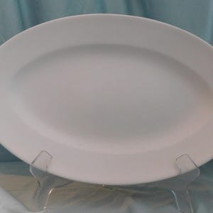 May include: A plain white oval serving platter with a smooth, matte finish and a wide, slightly raised rim. The platter is displayed on a clear plastic stand, showcasing its simple, clean, and functional design for food presentation.