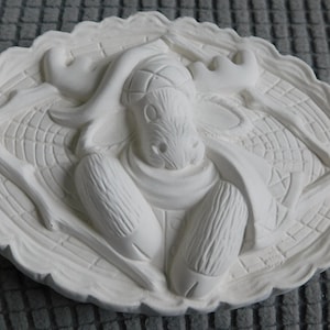 May include: A white, oval-shaped relief sculpture featuring a moose. The moose has large antlers and is surrounded by a decorative border. The sculpture is made of a solid, smooth material.