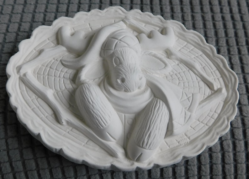 May include: A white, oval-shaped relief sculpture featuring a moose. The moose has large antlers and is surrounded by a decorative border. The sculpture is made of a solid, smooth material.