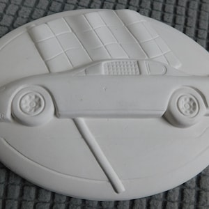 May include: A white oval-shaped ceramic plaque featuring a race car design. The car is depicted in profile, with a checkered flag in the background. The plaque is a solid, unpainted piece, suitable for painting or other customization.