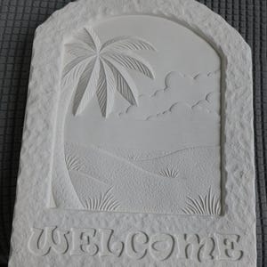 May include: A white stone welcome sign with a carved tropical scene. The design features a palm tree, rolling hills, and the word "WELCOME" at the bottom. The sign has a textured, rough-hewn appearance.