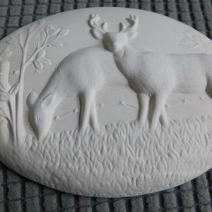 May include: White oval relief sculpture featuring two deer in a grassy field with a tree and fence. One deer is grazing, while the other stands close by. The artwork is a monochromatic, detailed depiction of wildlife.