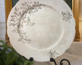 May include: A vintage white plate with a scalloped edge, adorned with a floral design in shades of gray and brown. The plate is displayed on a black metal stand, with a decorative lamb figurine in the foreground. The plate measures approximately 10 inches in diameter.