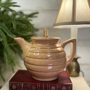 May include: A tan ceramic teapot with horizontal ribbed detailing, a curved handle, and a small lid handle. The teapot sits atop a vintage burgundy book. A lamp and decorative pineapple are in the background.
