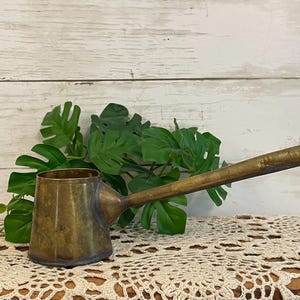 May include: A vintage brass coffee pot with a long, slender handle and a cylindrical body. The metal surface shows a weathered, aged patina with visible dark spots and metallic texture, featuring a wide opening at the top and a tapered base.