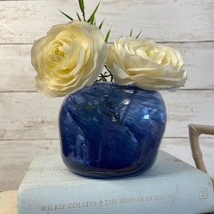May include: A blue glass vase with a swirling pattern holds two large, creamy white roses and green stems. The vase sits atop a light blue book titled "WILKIE COLLINS THE WOMAN IN WHITE". The background is a white wooden wall.