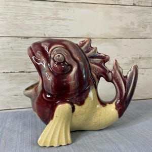 May include: A ceramic fish-shaped pitcher featuring a glossy glaze with deep burgundy and purple tones on the upper body and head, transitioning to a cream-colored base with a visible crackle finish on the fins and lower body sections.