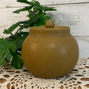May include: A round, mustard-yellow ceramic jar with a smooth, matte finish and a matching lid featuring a small, spherical knob handle. The container has a wide, bulbous body that tapers slightly toward the top opening, presenting a simple, solid-colored design.