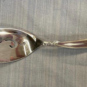 May include: Silver-colored serving spoon with a slotted, oval head and a decorative handle. The spoon features a curved design with ornate detailing near the handle's end. The slotted head has three decorative cutouts.