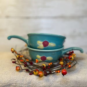 May include: Two turquoise ceramic bowls with handles are stacked, each adorned with a floral design in yellow and red. A decorative garland of orange, yellow, and red berries encircles the bowls, creating a rustic, autumnal aesthetic.