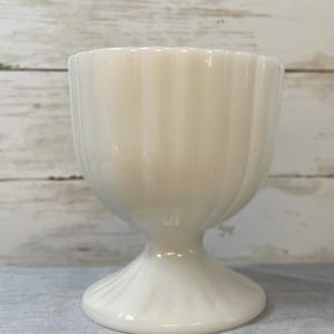 May include: A white, vintage-style egg cup with a fluted design. The egg cup has a pedestal base and a rounded bowl. The color is a creamy white, and the surface has a glossy finish. The egg cup is made of ceramic or porcelain.