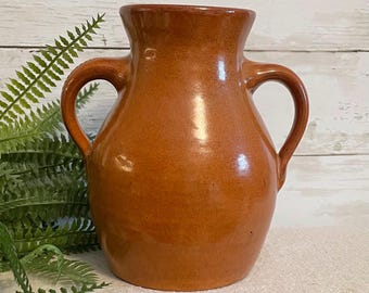 May include: A glossy, burnt-orange ceramic vase featuring a wide, rounded body, a flared rim, and two symmetrical curved handles on opposite sides. The surface has a smooth, reflective finish with subtle variations in the warm, earthy color tone.