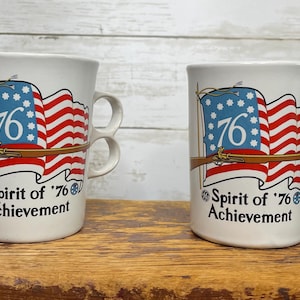 May include: Two white ceramic mugs with a patriotic design. Each mug features a depiction of the American flag with the number '76' and the text 'Spirit of '76 Achievement'. The mugs have a unique handle design.