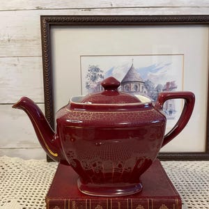 May include: A burgundy ceramic teapot with a gold crackle pattern and a matching lid. The teapot sits on a red book with gold lettering. A framed picture is in the background.